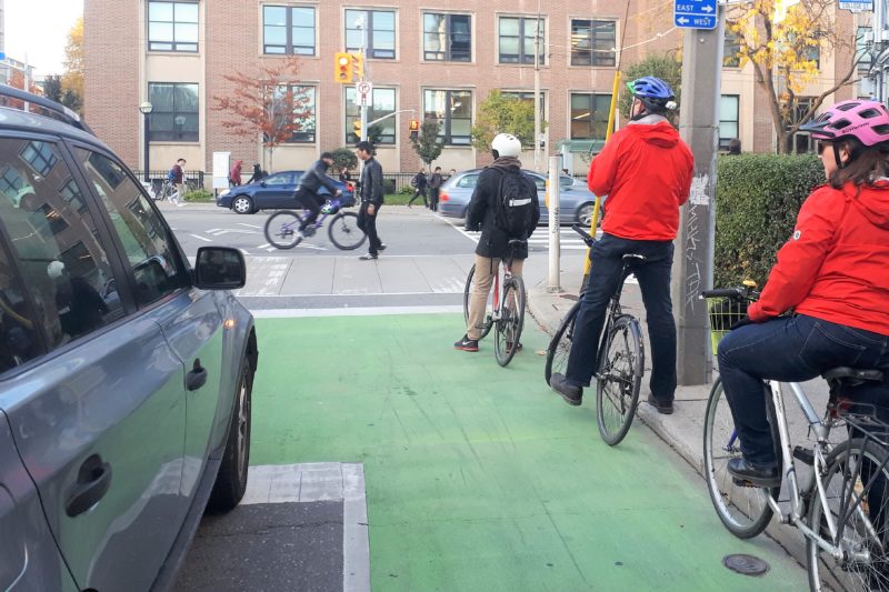 Three cyclists wait at a light in a green, painted bike box with a car beside them